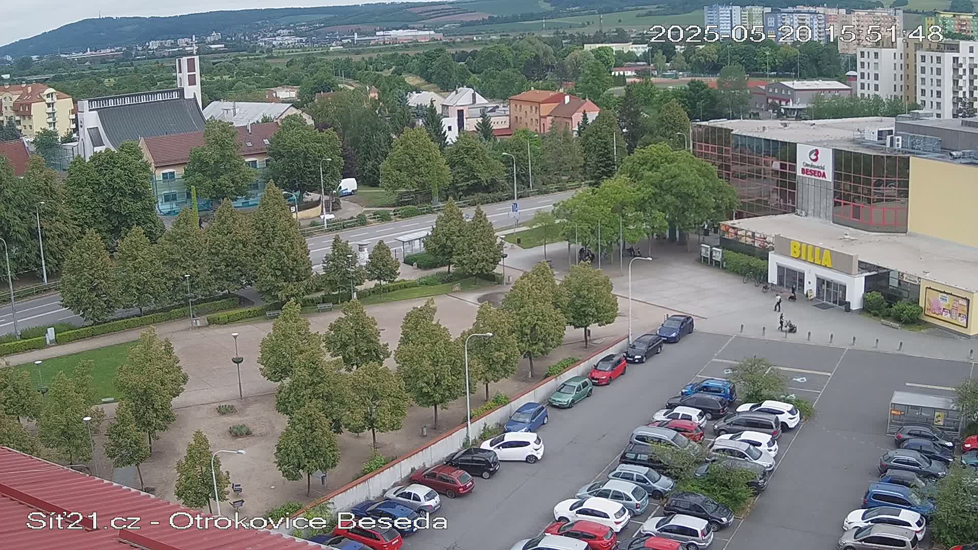 An aerial view shows a town square with a church, a large supermarket, and a parking lot filled with cars on an overcast day.