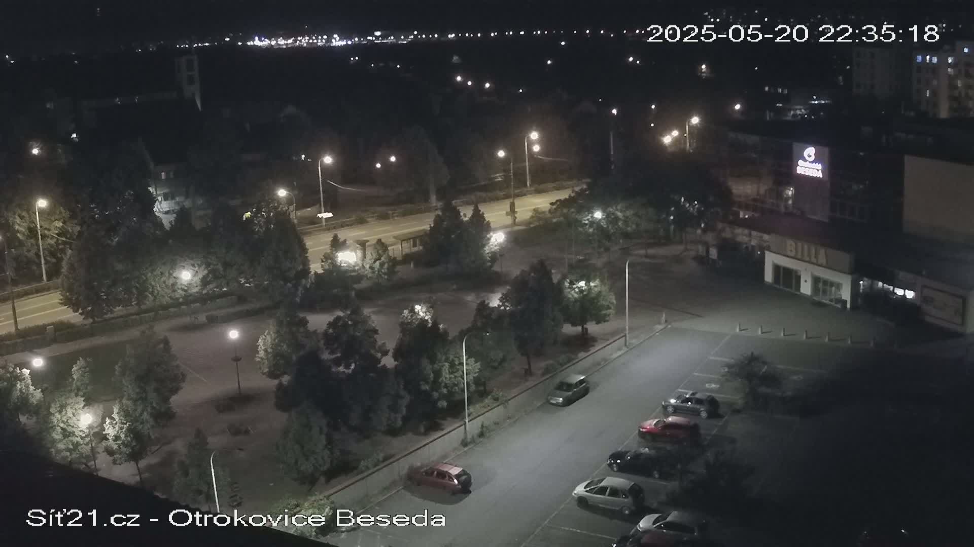 A nighttime, high-angle view shows a parking lot with several cars, surrounded by trees and streetlights, next to a road and a building.