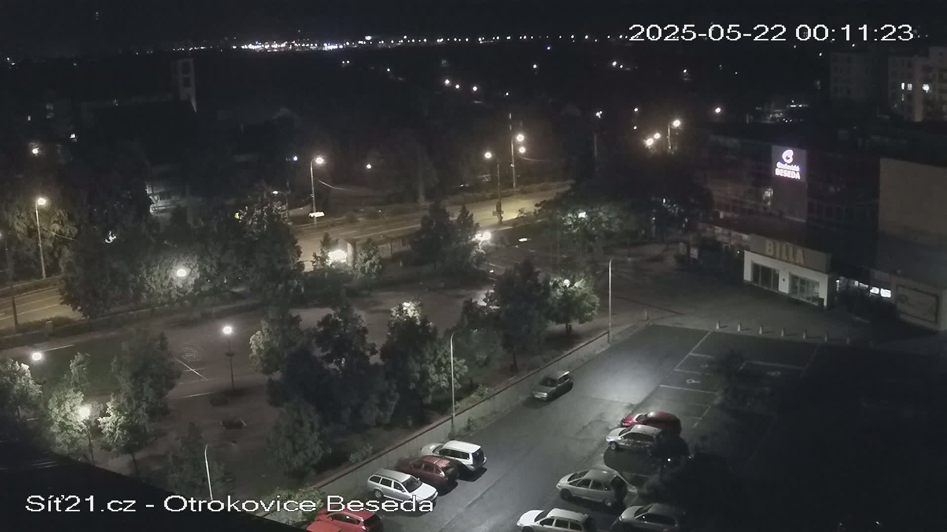 A nighttime, high-angle view of a parking lot with several cars, surrounded by trees and streetlights, near a road and buildings.