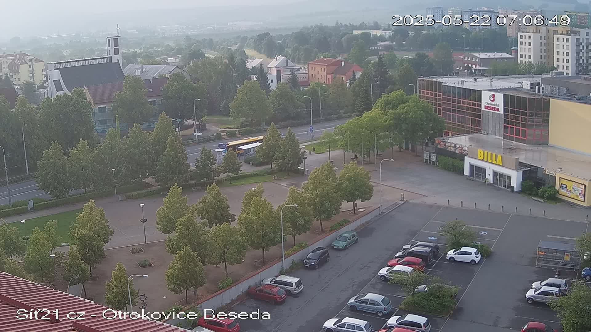 A hazy view of a town square with a church, a supermarket, and a parking lot containing several cars.