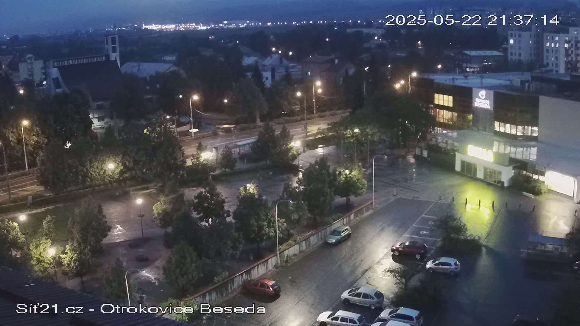 A nighttime aerial view of a town, showing wet streets, cars in parking lots, and buildings with illuminated lights under a dark, overcast sky.