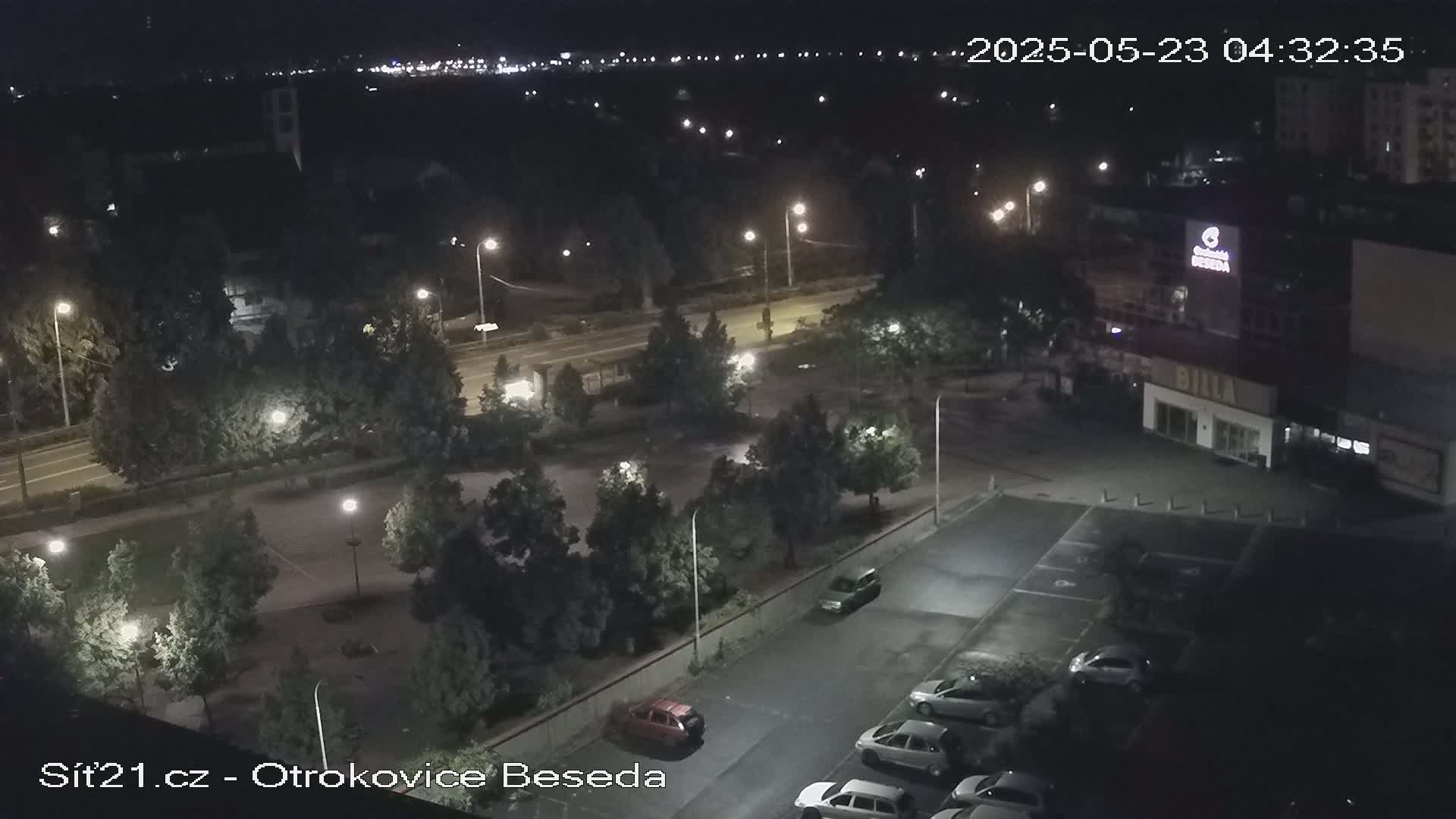 A nighttime, high-angle view of a parking lot and roadway, illuminated by streetlights, with several cars parked and a few trees surrounding the area.