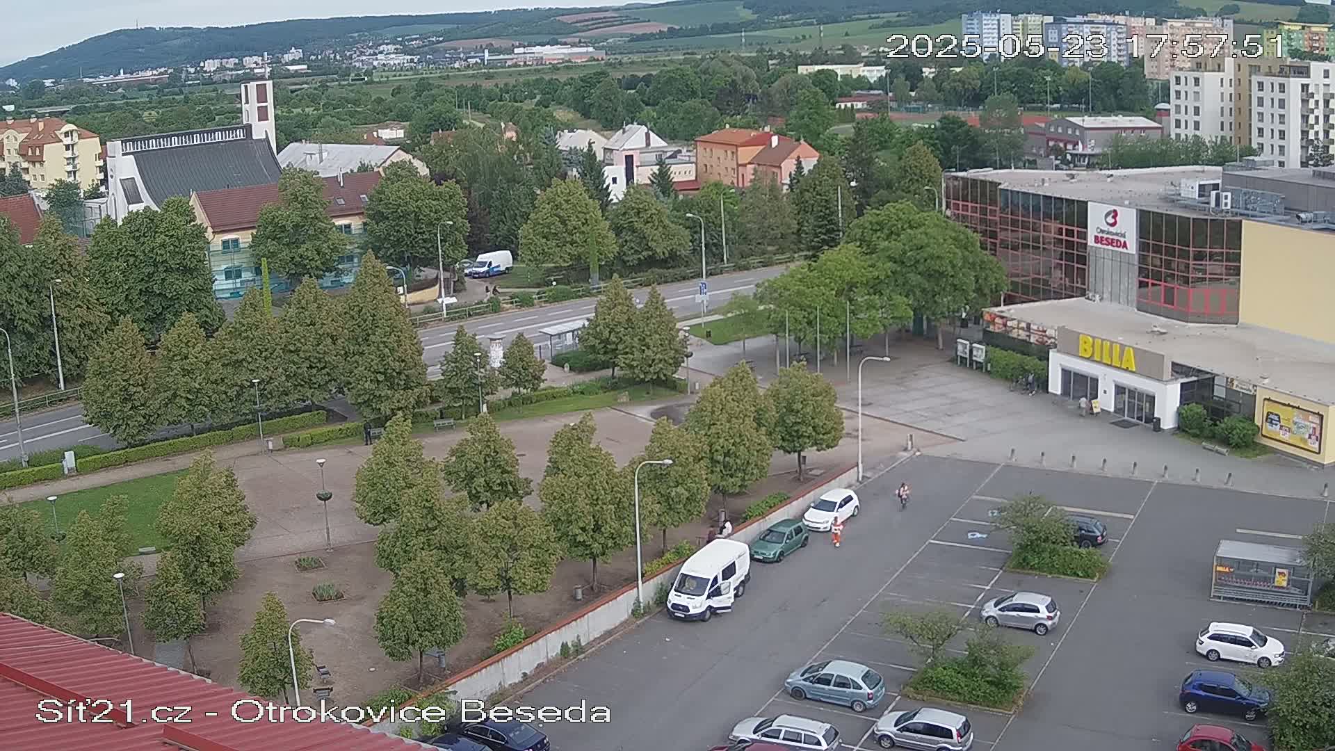 An aerial view shows a parking lot, a supermarket, a church, and a town square with several cars and pedestrians, under a partly cloudy sky.