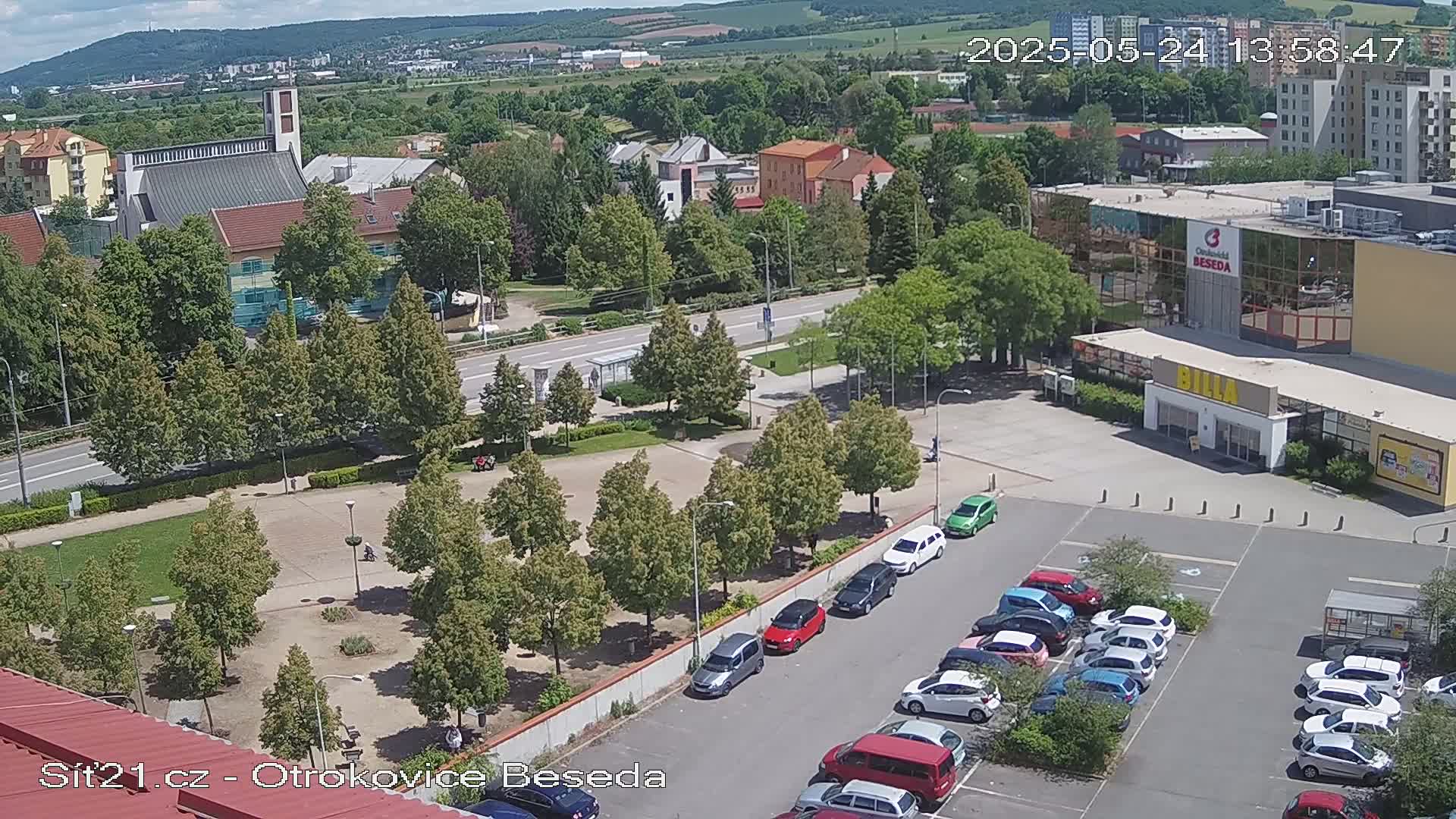 A high-angle, sunny view of a town square with a church in the background, green spaces, and a parking lot full of cars.