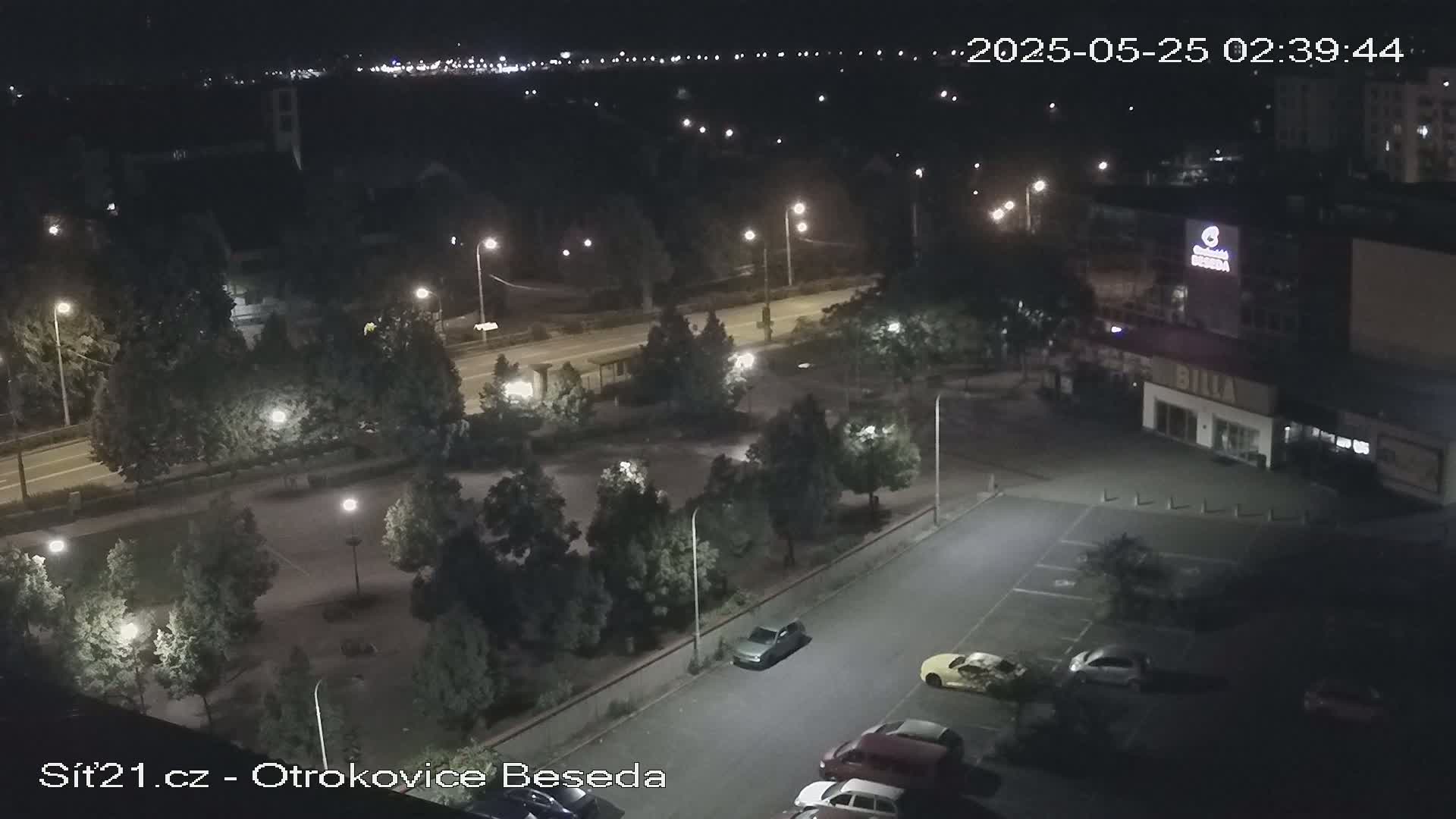 A nighttime aerial view of a parking lot, a road, and a park-like area with trees and streetlights, showing several cars parked.