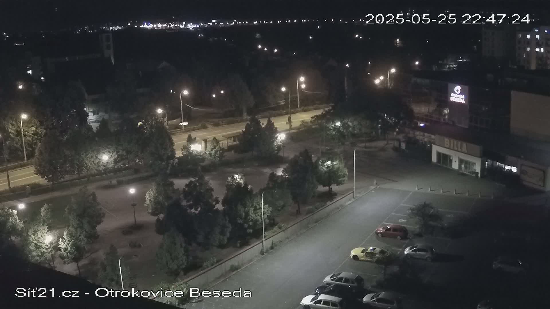 A nighttime aerial view of a parking lot and park-like area next to a road, with several cars parked and streetlights illuminating the scene.