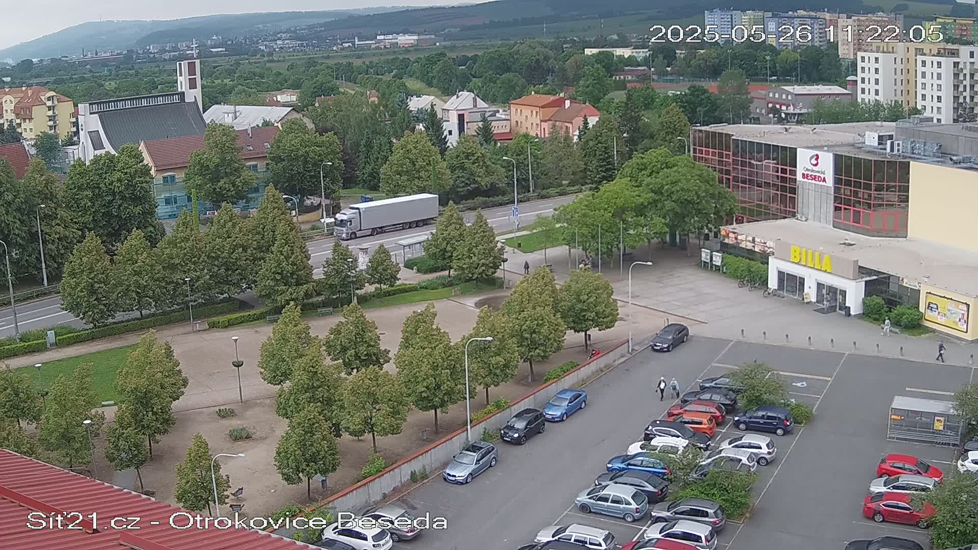 An overcast day reveals a town square with a parking lot full of cars, a church in the background, and a large building with a supermarket.