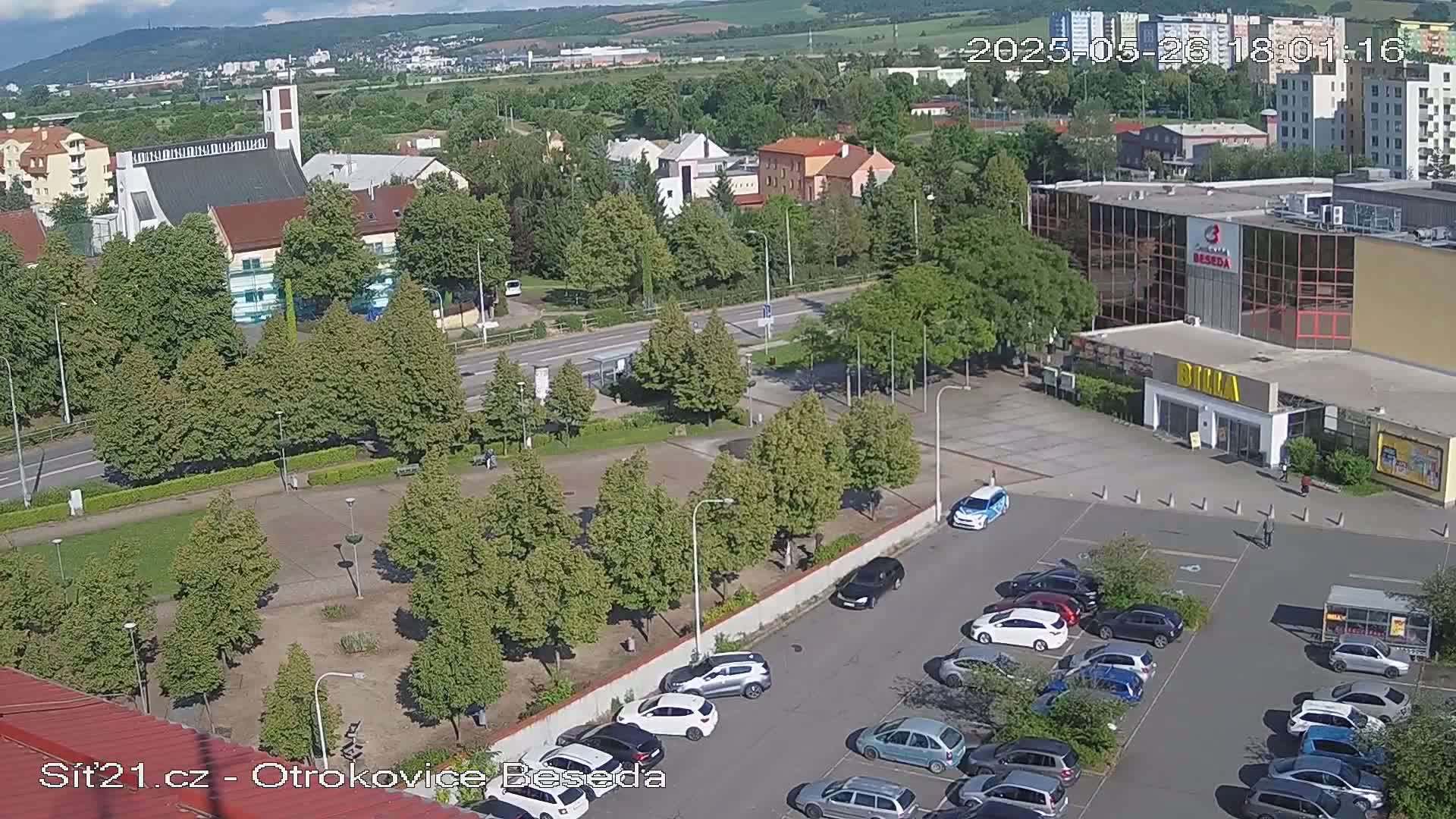 A high-angle view shows a town square with a church, buildings, and a parking lot full of cars on a sunny day.