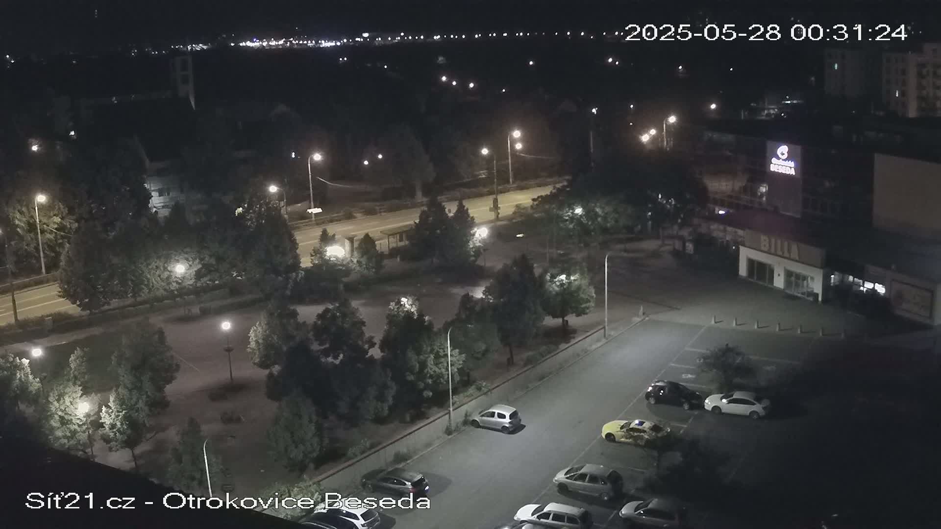 A nighttime aerial view of a parking lot and roadway, surrounded by trees and buildings, illuminated by streetlights.