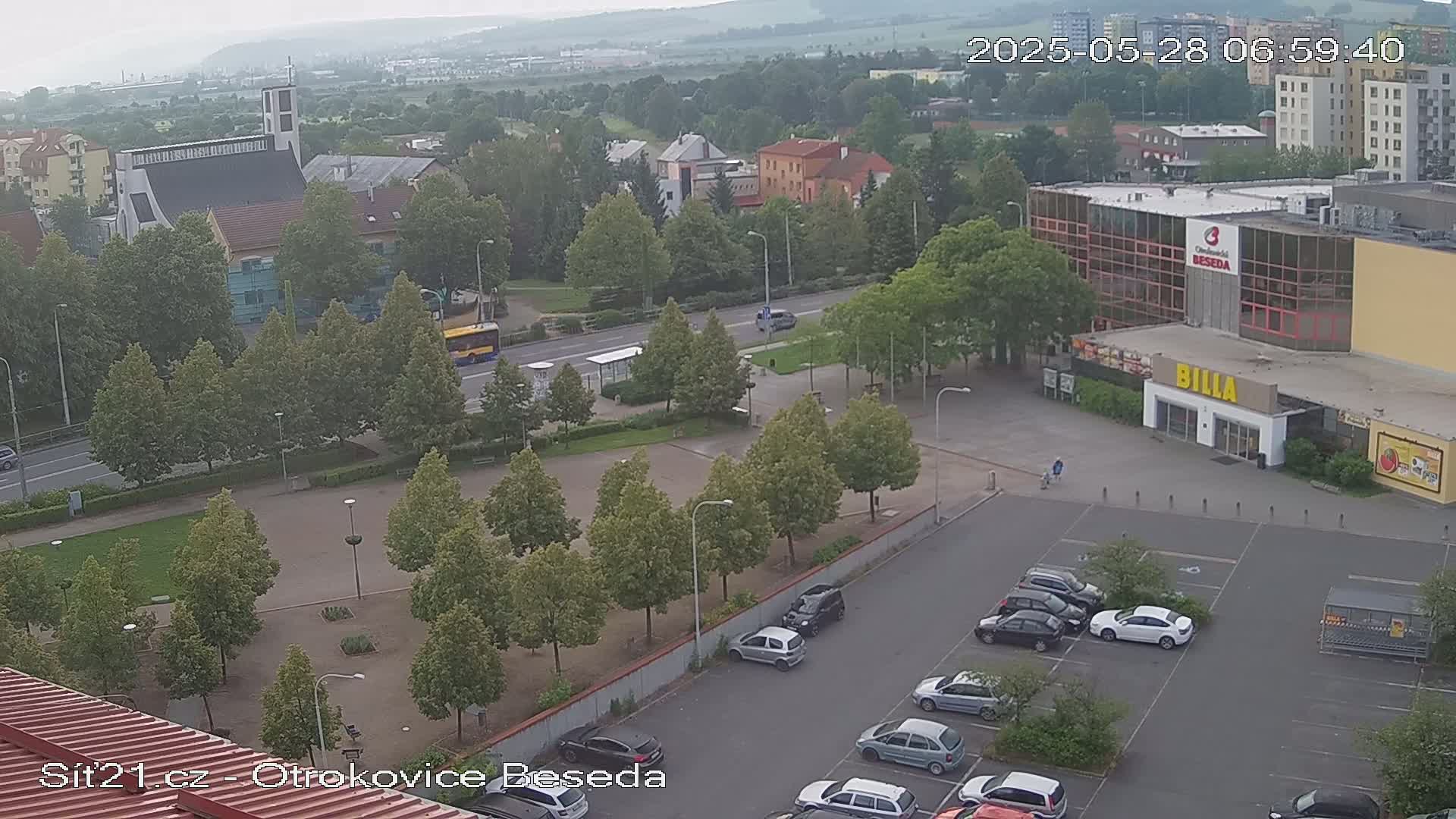 A high-angle, long shot on an overcast day shows a town square with a church, a large building, a parking lot, and a bus, all surrounded by trees.