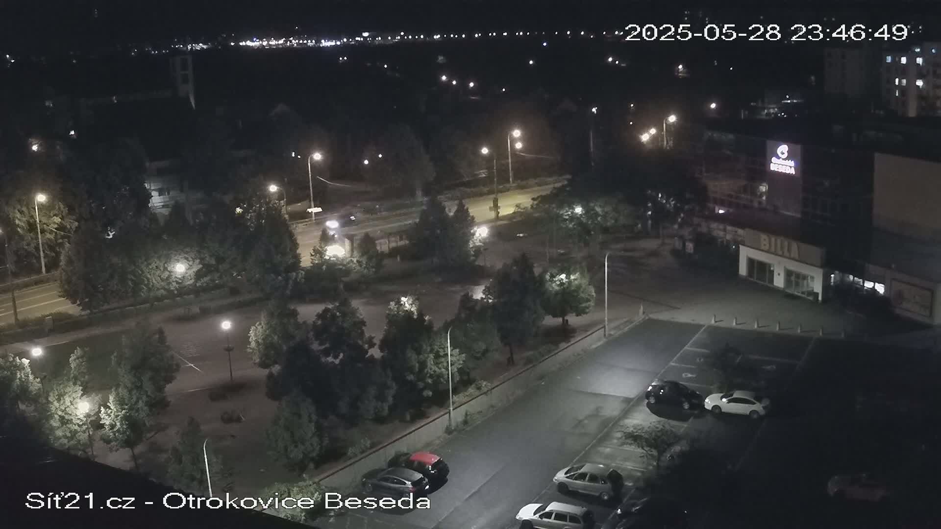 An elevated nighttime view of a parking lot and roadway, illuminated by streetlights, with several cars parked and a few trees scattered throughout.