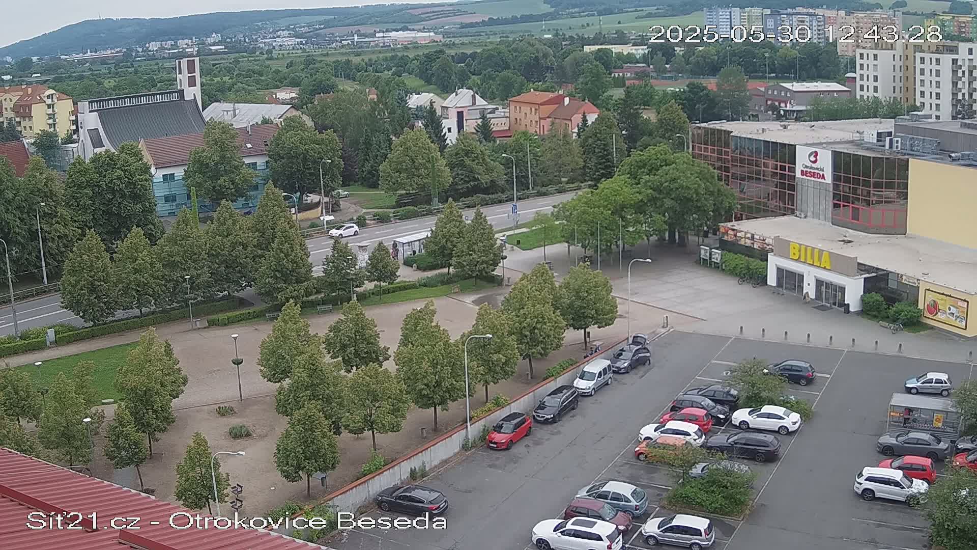 An aerial view of a town square with a church, a large building, and a parking lot filled with cars on an overcast day.