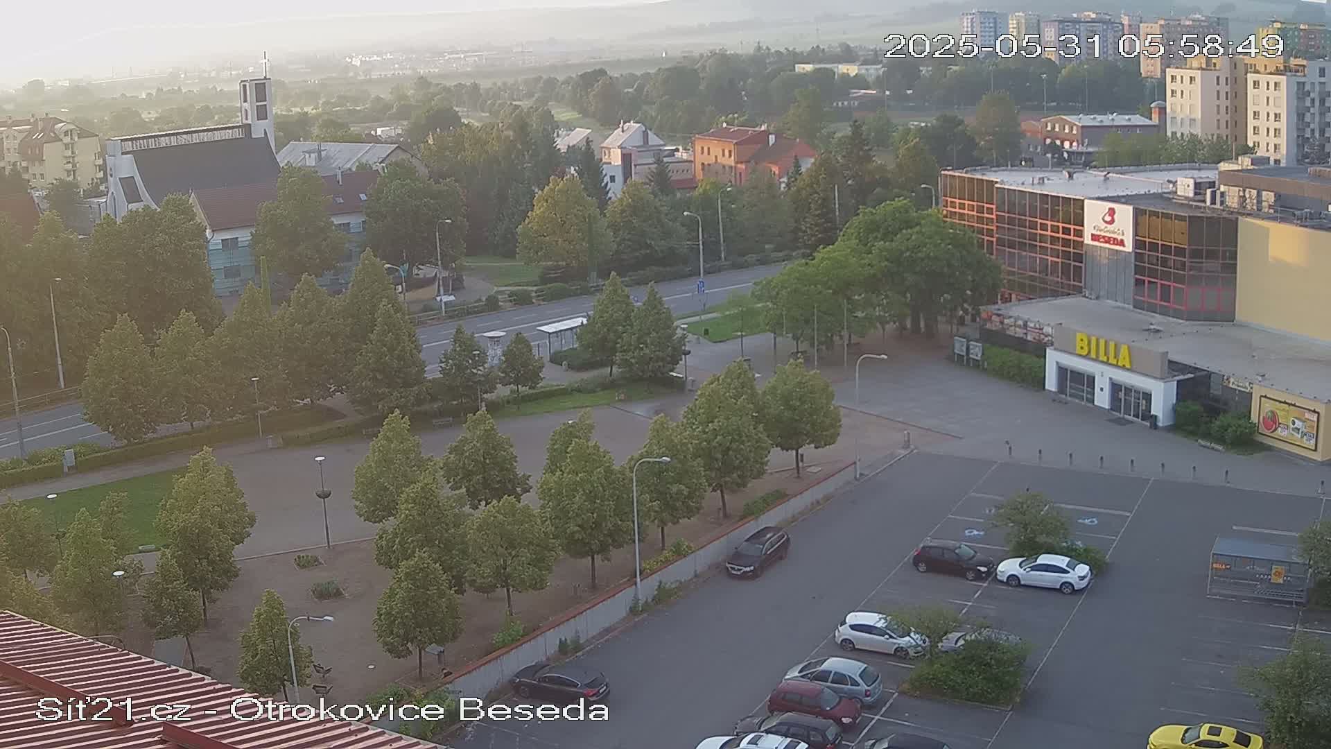 A hazy, high-angle view shows a town square with a church, buildings, and a parking lot containing several cars, all surrounded by lush green trees.