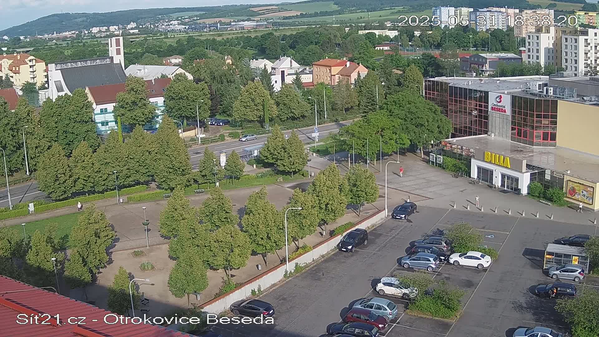 An aerial view of a town square on a sunny day, showing a church, shops, a parking lot with cars, and a grassy area with trees.