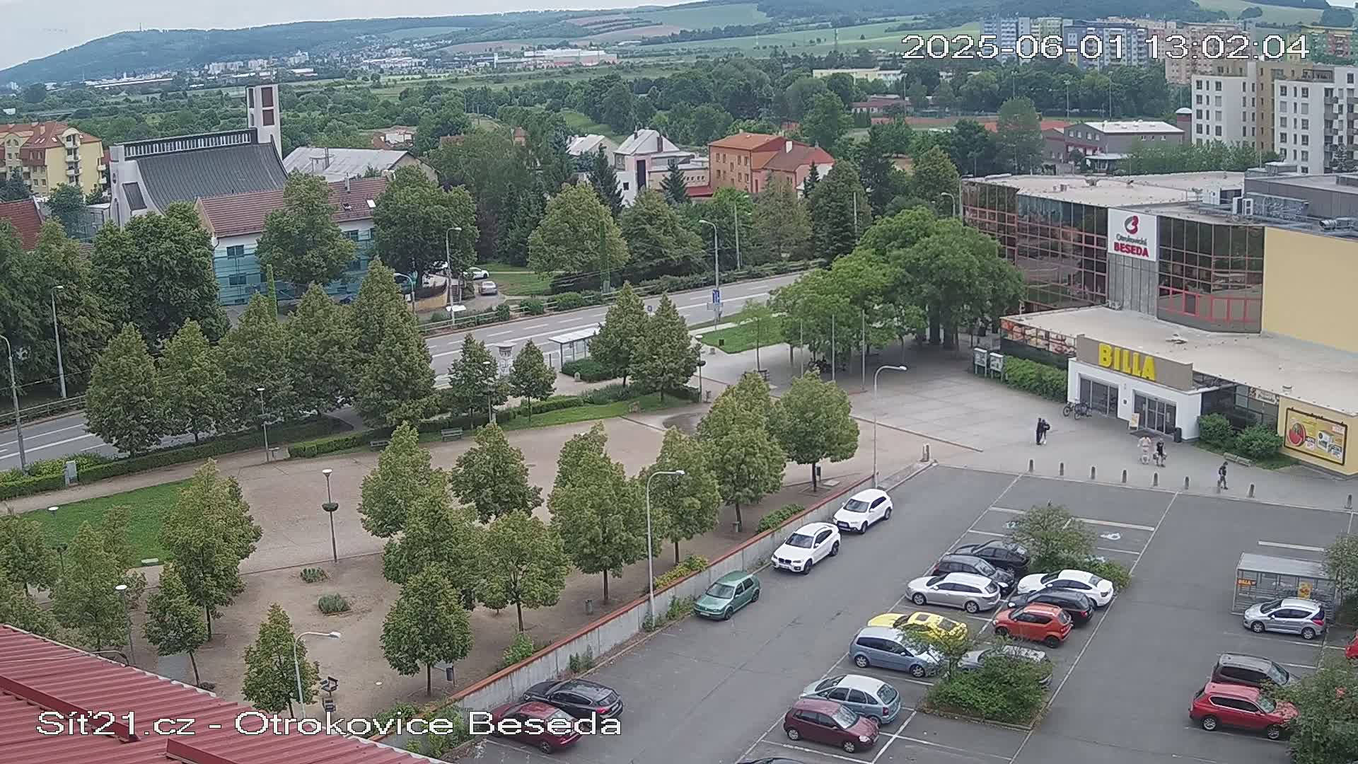 An aerial view shows a town square with a church, a supermarket, and several cars parked in a lot, under a partly cloudy sky.
