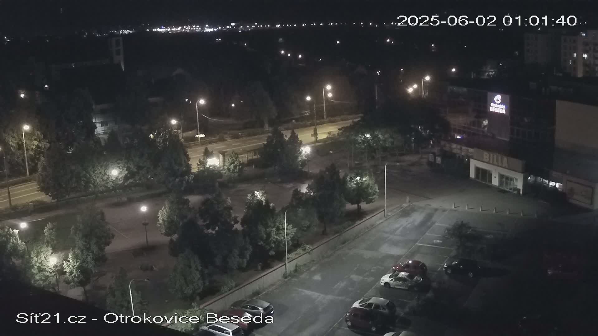 A nighttime, overhead view of a parking lot with several cars, a road with streetlights, and trees surrounding a small park-like area next to a building.