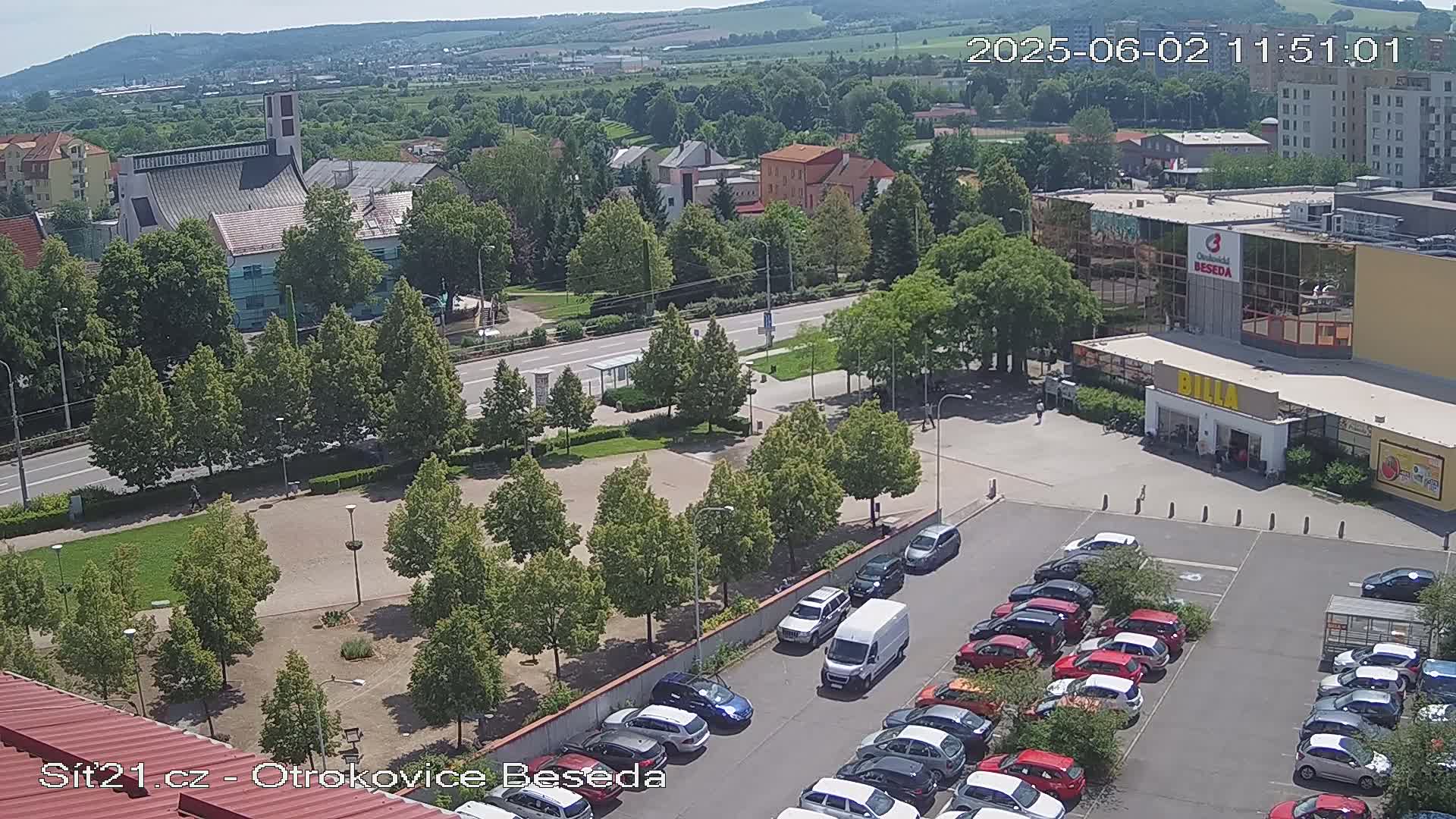 A high-angle, sunny view shows a town square with a church and a parking lot filled with cars next to a shopping center.