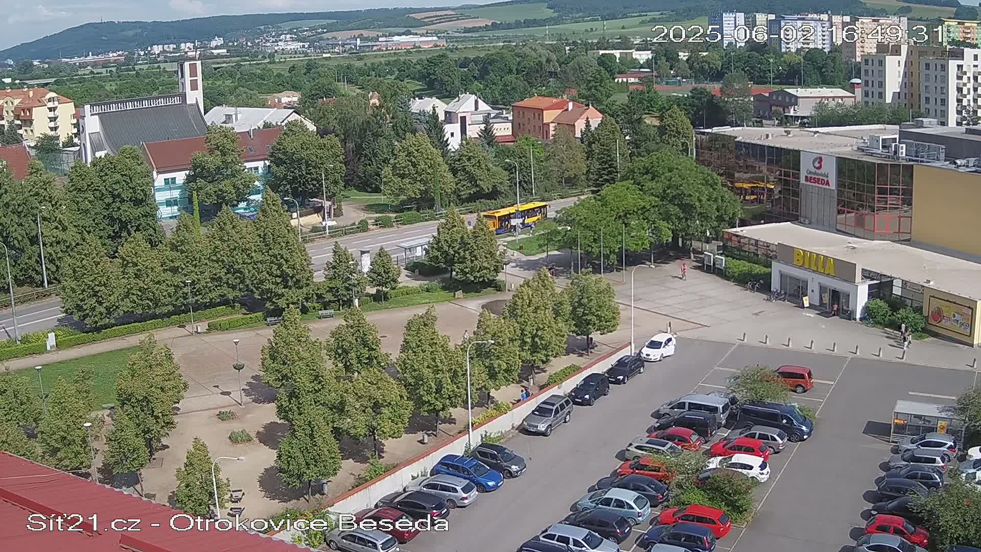 An aerial view shows a town square with a church, a bus, and a parking lot full of cars on a sunny day.