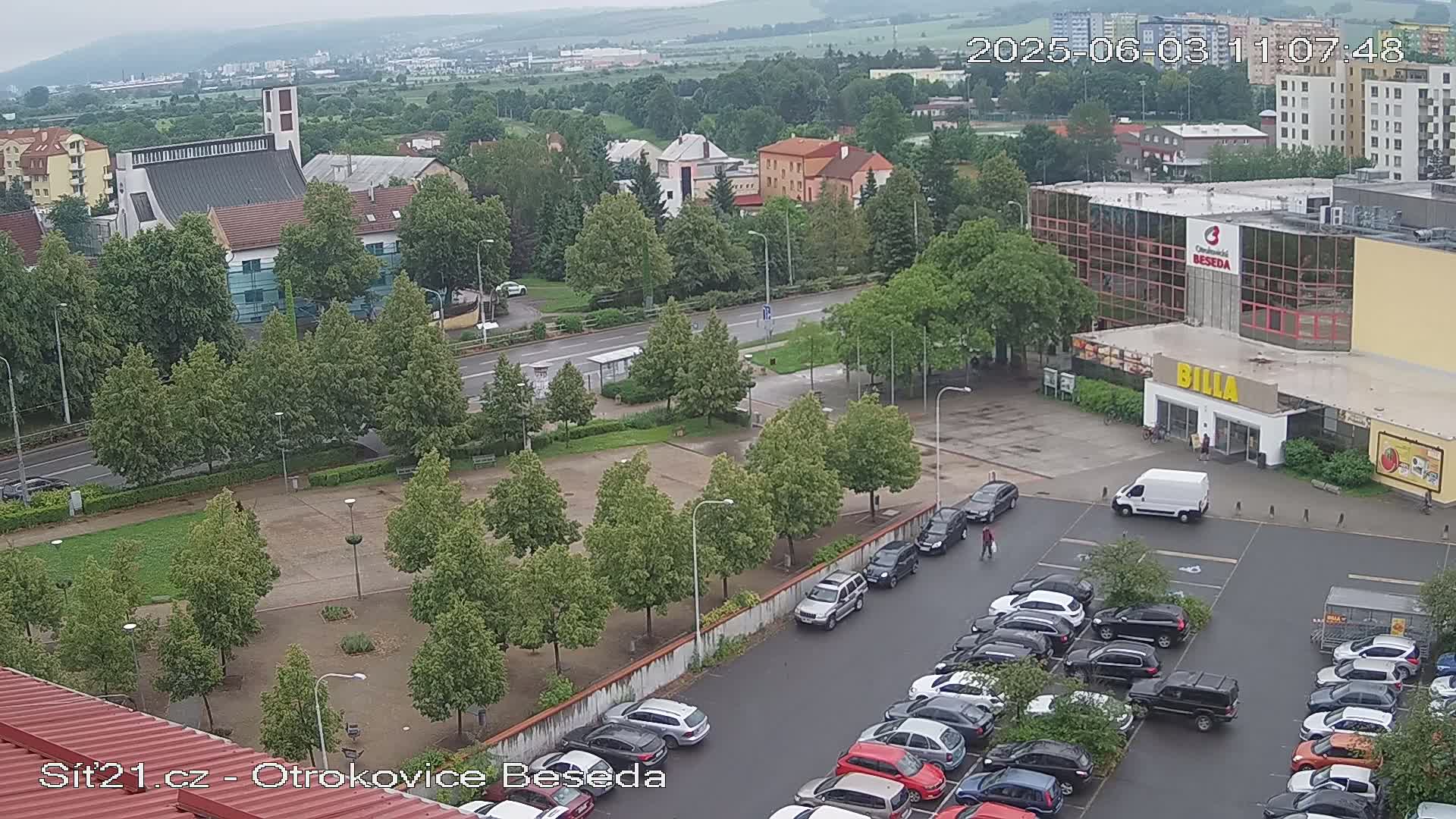 An overcast day reveals a parking lot full of cars next to a shopping center and church, surrounded by green trees and a paved area.