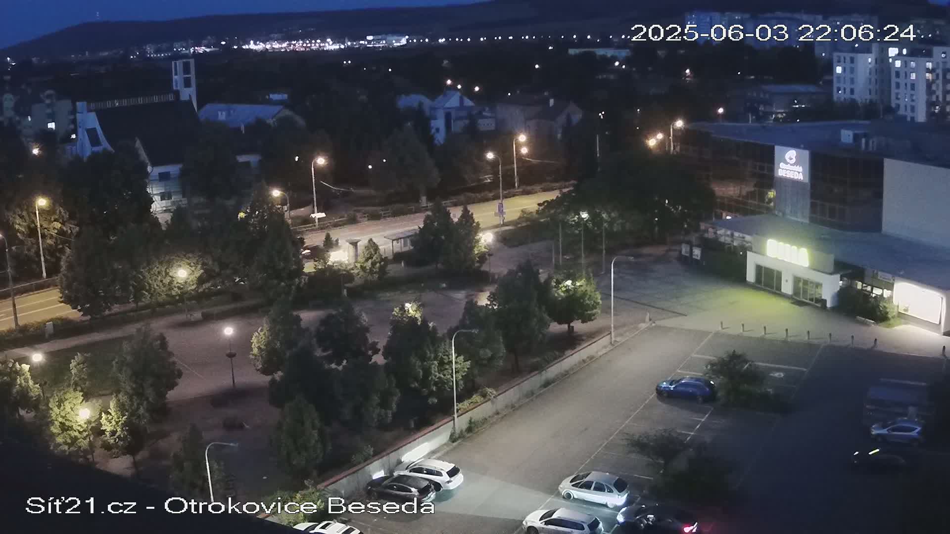 A nighttime aerial view of a parking lot with several cars, streetlights illuminating a roadway and surrounding trees, and a building in the background under a clear night sky.