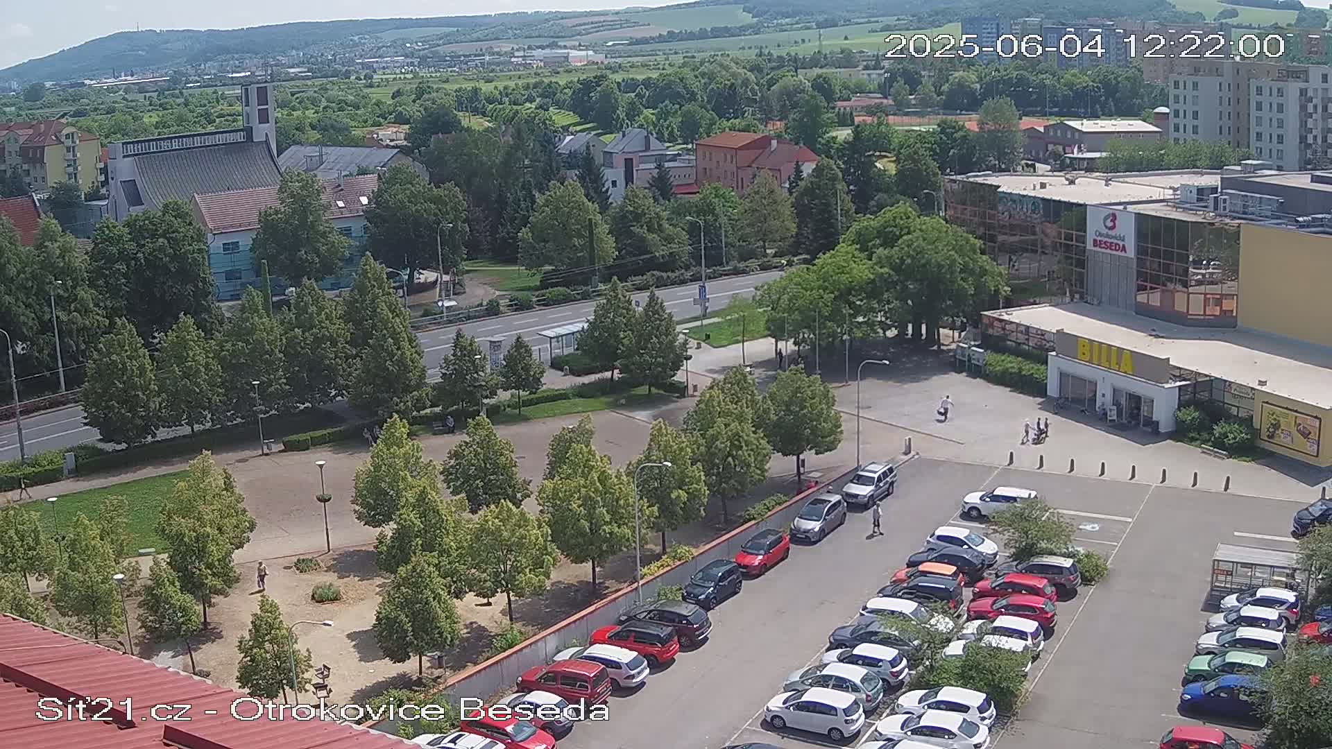 An aerial view of a town square with a parking lot full of cars, a church, and several buildings on a sunny day.