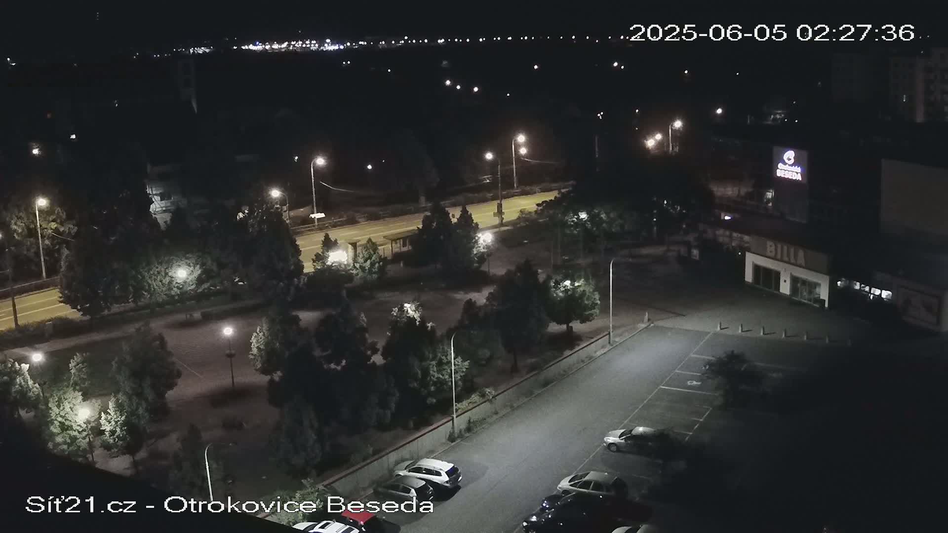 An aerial nighttime view of a parking lot with several cars, a road with streetlights, and a building with illuminated signage.