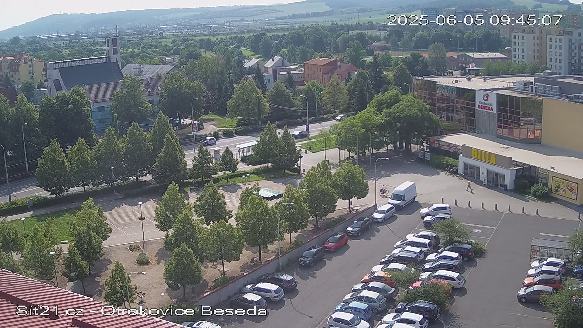 A high-angle, sunny day view of a town square with a large parking lot full of cars, a church, and a shopping center.
