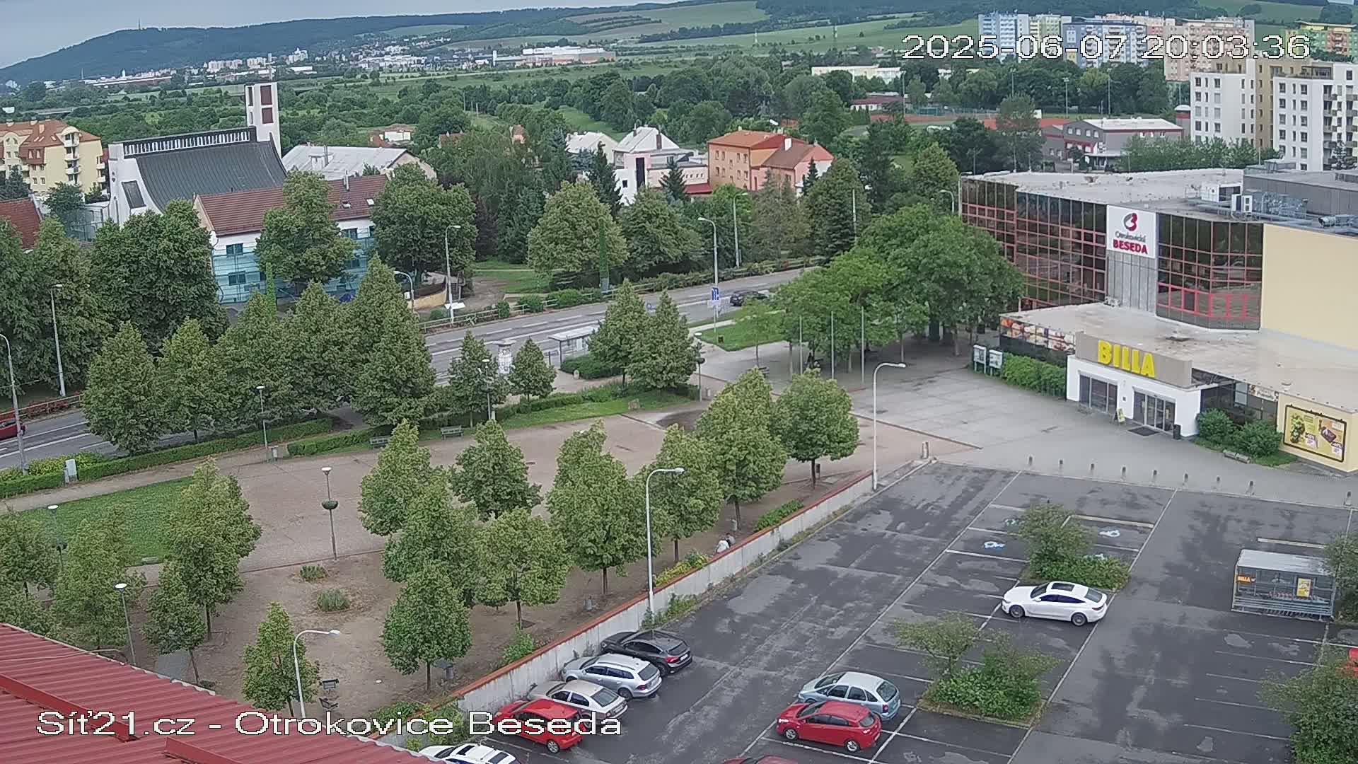 An aerial view shows a town square with a church, buildings, a parking lot with cars, and green spaces, under an overcast sky.