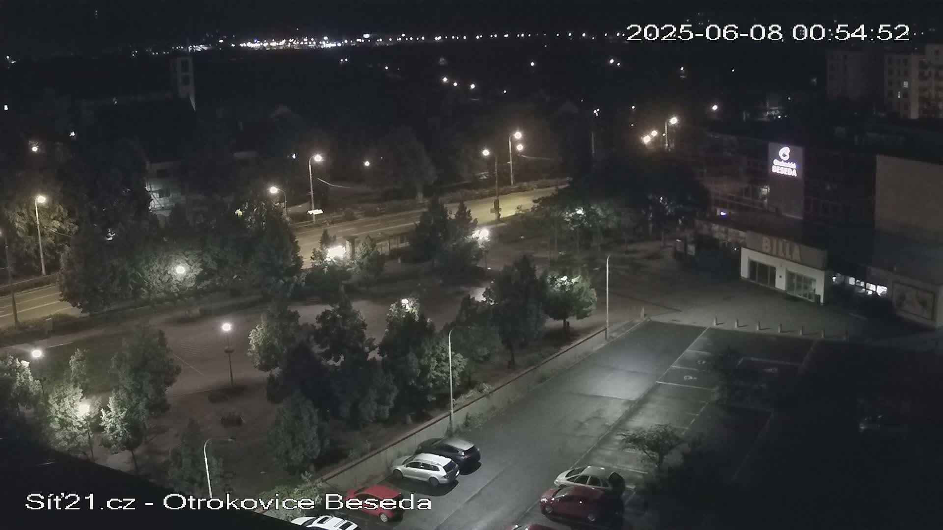 A nighttime aerial view of a parking lot and road, illuminated by streetlights, with several cars parked and trees surrounding the area.
