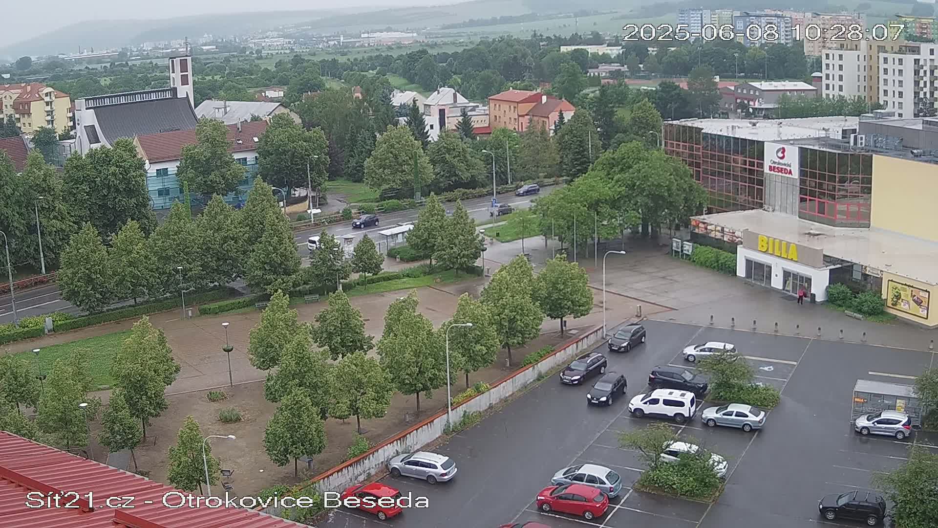 An elevated view of a town square on a cloudy day shows a church, buildings, cars in a parking lot, and a large building with a supermarket.