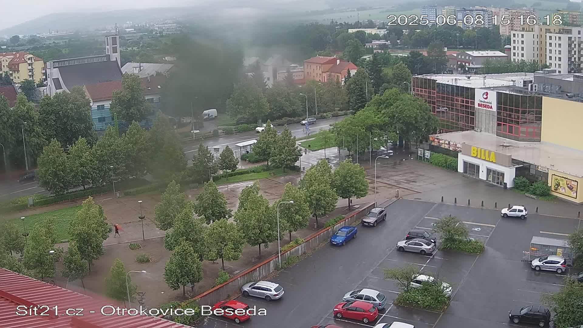 A rain-soaked town square with a church in the background, cars parked in a lot, and several trees lining a walkway, all under an overcast sky.