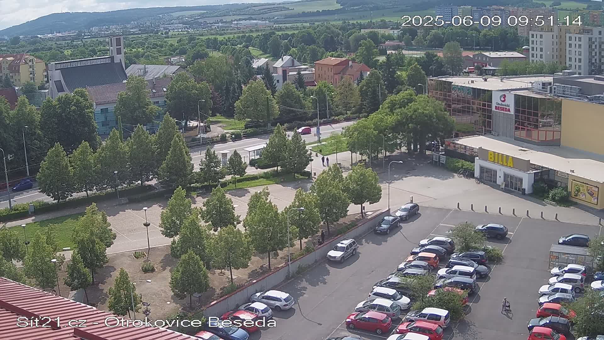 A high-angle, sunny-day view of a town square with a parking lot filled with cars, a church, several buildings, and many green trees.
