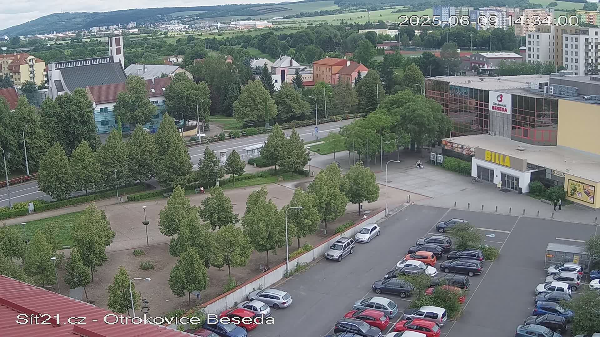 An aerial view of a town square with a church, buildings, a supermarket parking lot, and lush green trees under a partly cloudy sky.