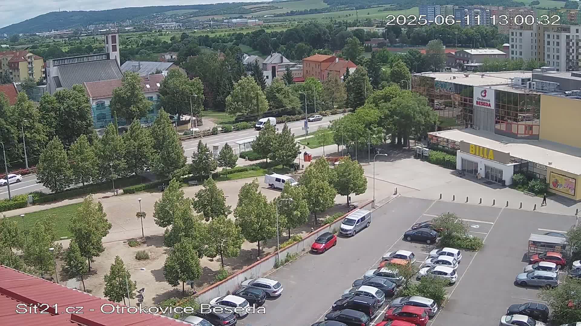 An aerial view of a town square on a sunny day, showing a church, shops, a parking lot filled with cars, and a grassy area with trees.
