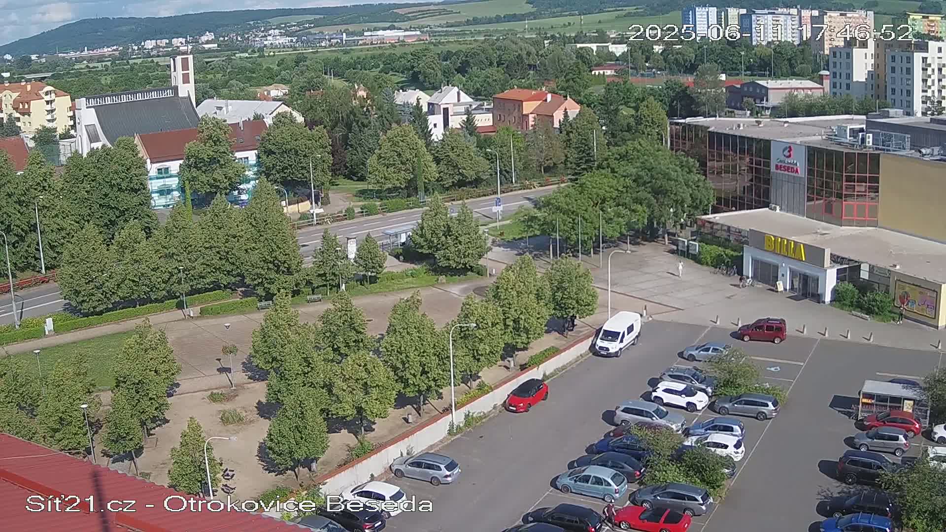 An aerial view of a town square on a sunny day shows a church, a supermarket, a parking lot full of cars, and a park with many green trees.