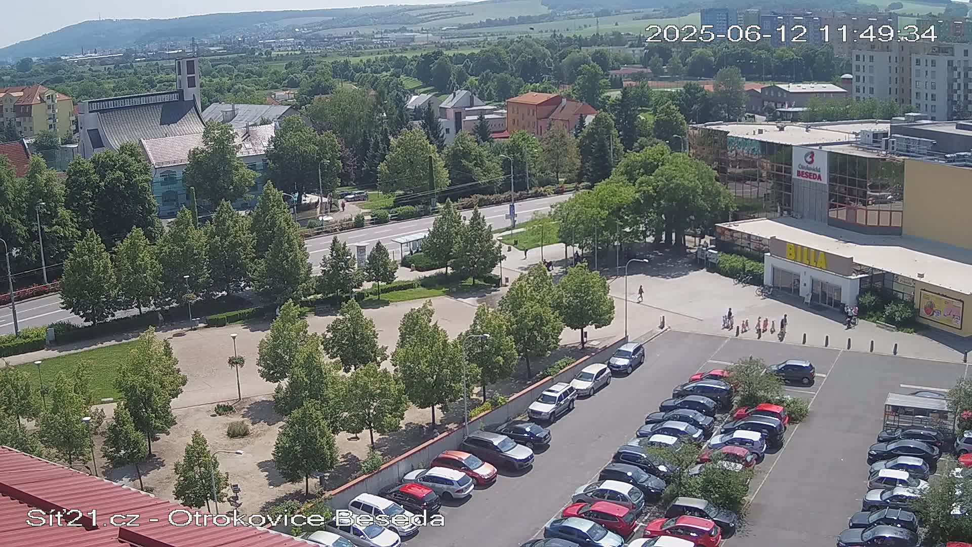 A high-angle, sunny view shows a town square with a church, a large parking lot filled with cars, and a supermarket, surrounded by green trees and buildings.