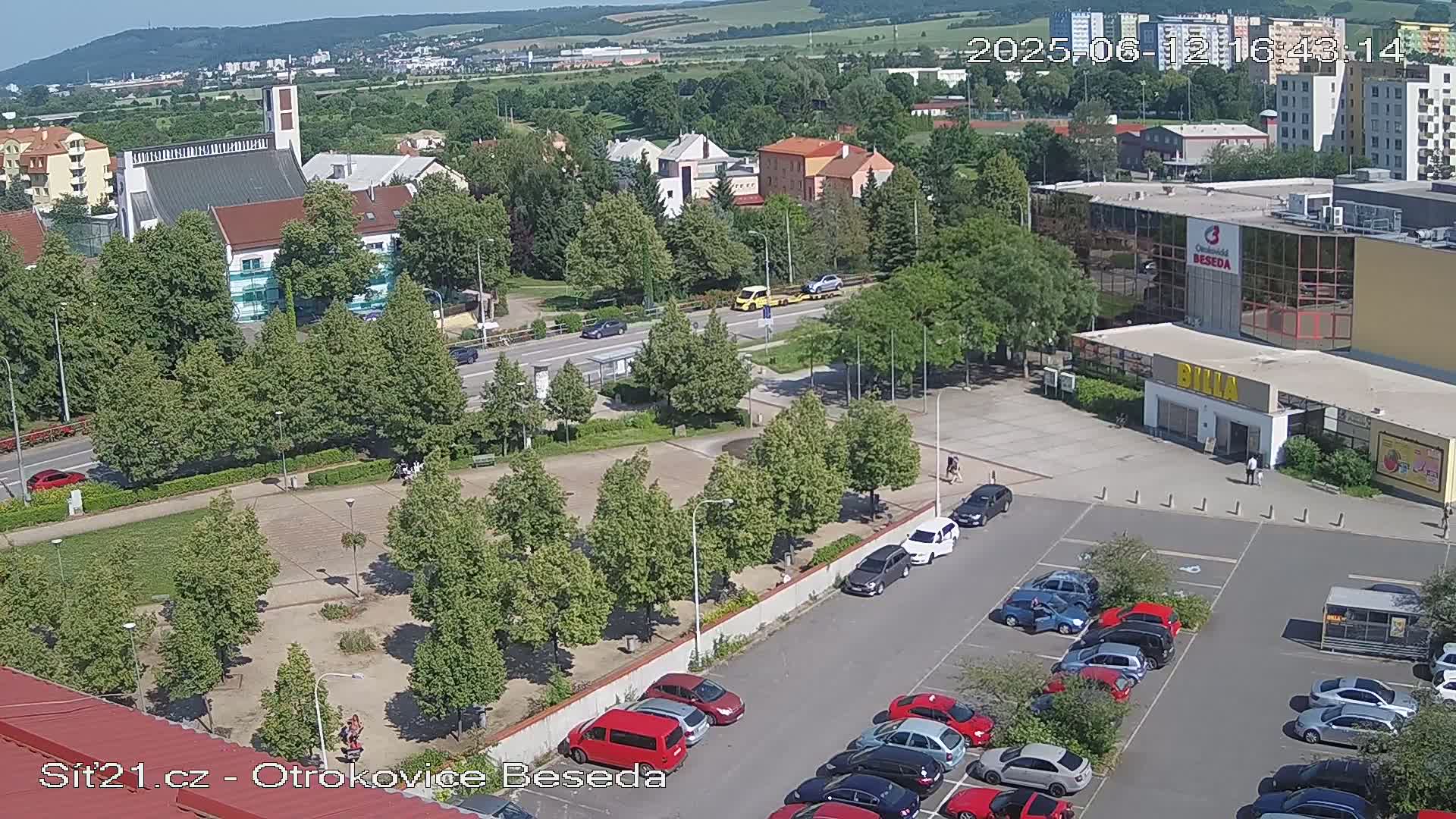 A high-angle view of a town square with a church, buildings, a parking lot full of cars, and green trees under a sunny sky.