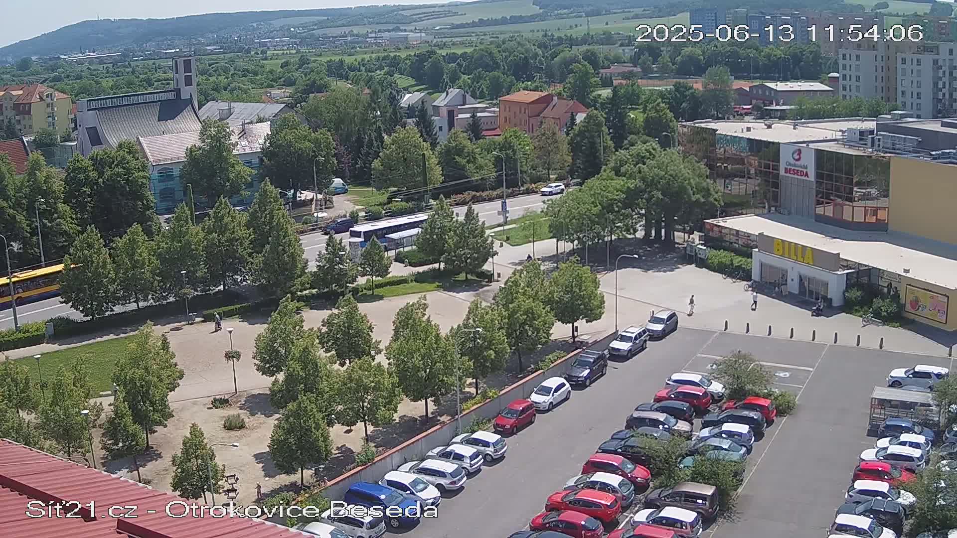 A sunny, overhead view of a town square with a church, a shopping center, and a full parking lot, surrounded by lush green trees.