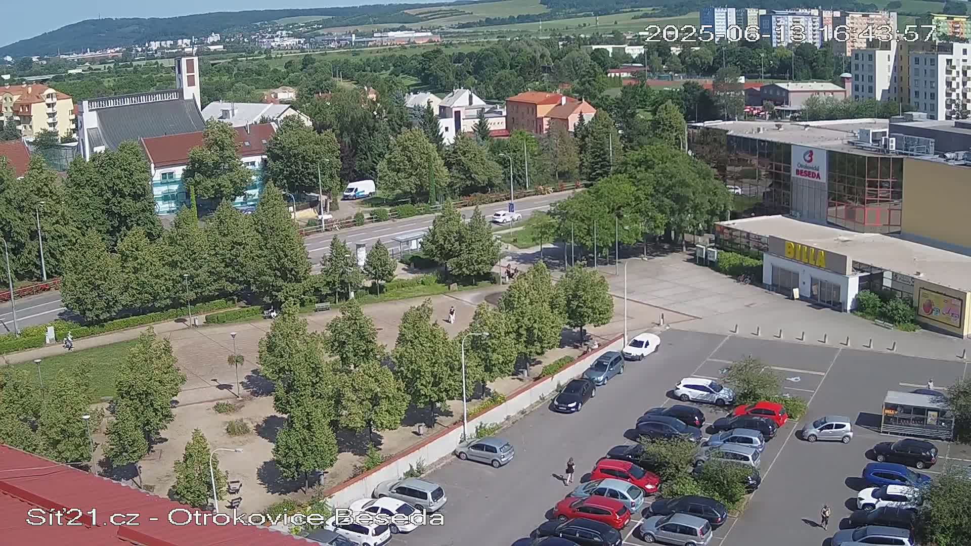 A high-angle view of a town square on a sunny day, showing a church, buildings, a parking lot filled with cars, and a park with trees.