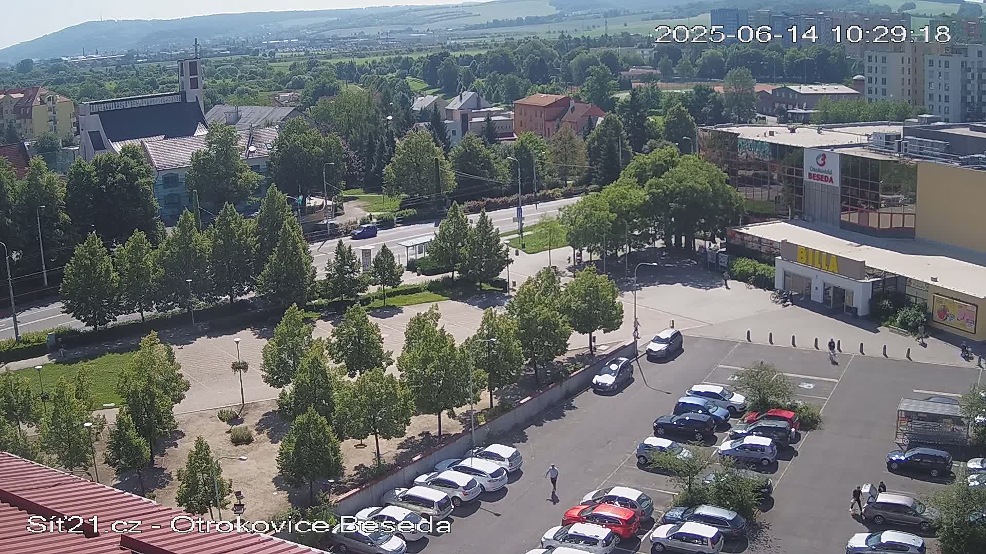 An aerial view shows a town square with a parking lot full of cars, a church, and a large building under a sunny sky.
