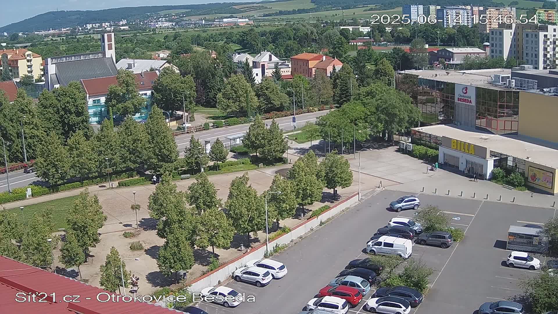 A sunny aerial view shows a town square with a church, buildings, a parking lot full of cars, and many green trees.