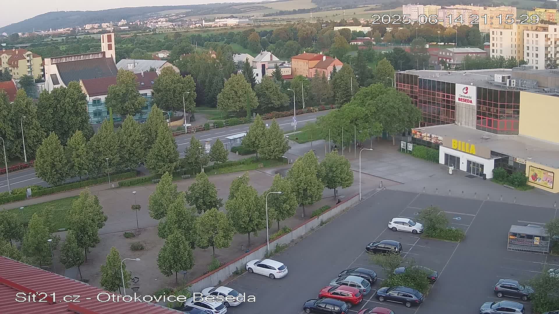 A sunny aerial view shows a town square with a church, buildings, a parking lot full of cars, and many green trees.
