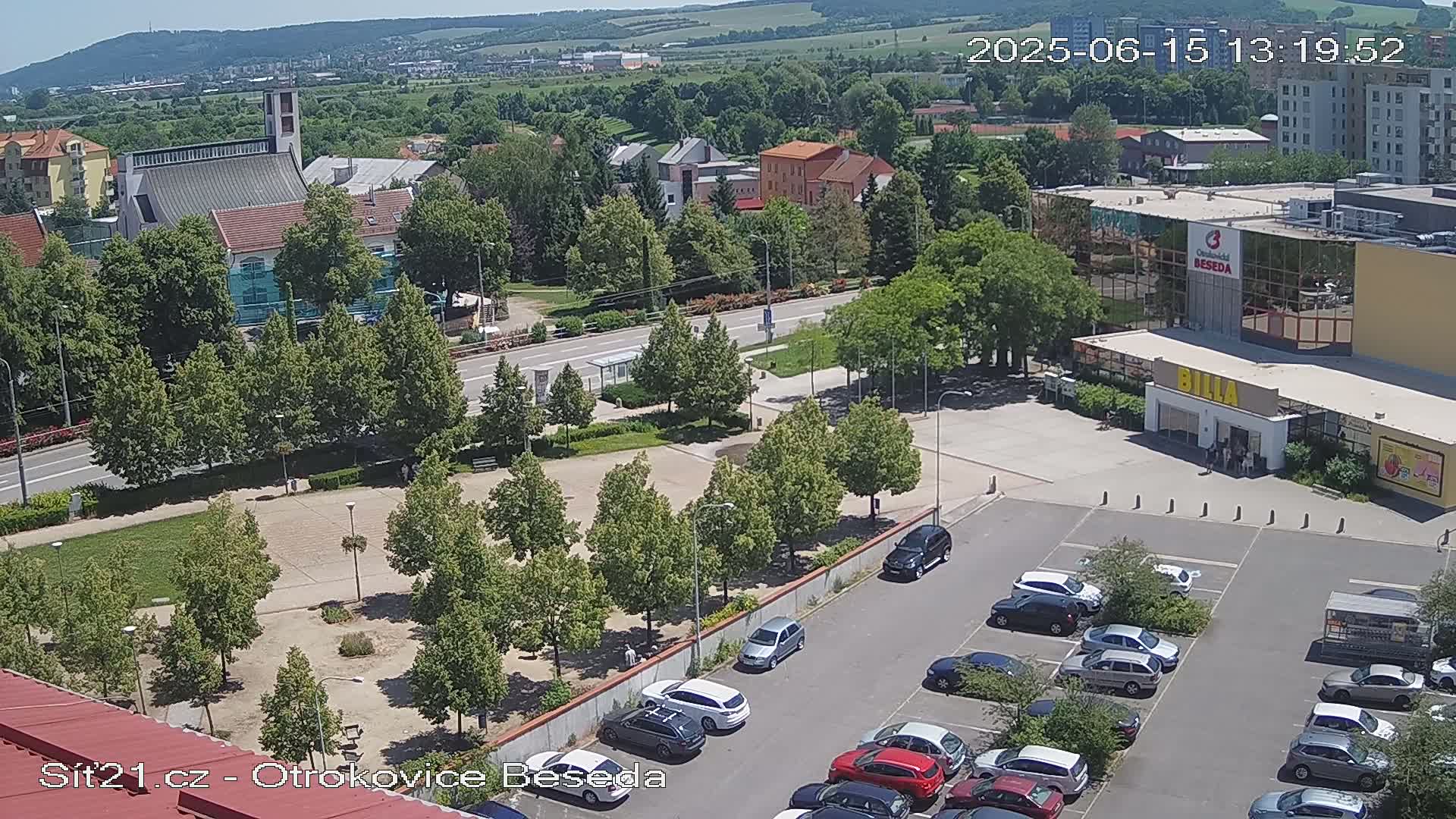 An aerial view on a sunny day shows a town square with a church in the background, a parking lot filled with cars, and a shopping center with a Billa supermarket.