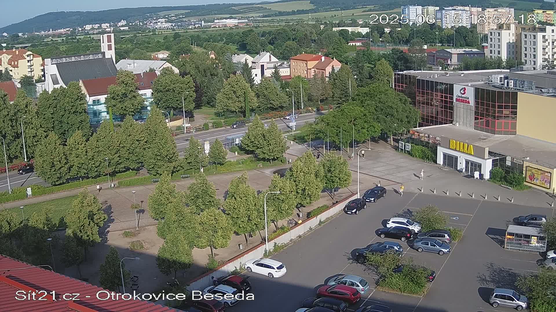 A high-angle, sunny view of a parking lot, a plaza with trees, buildings, and a road, all under clear skies.