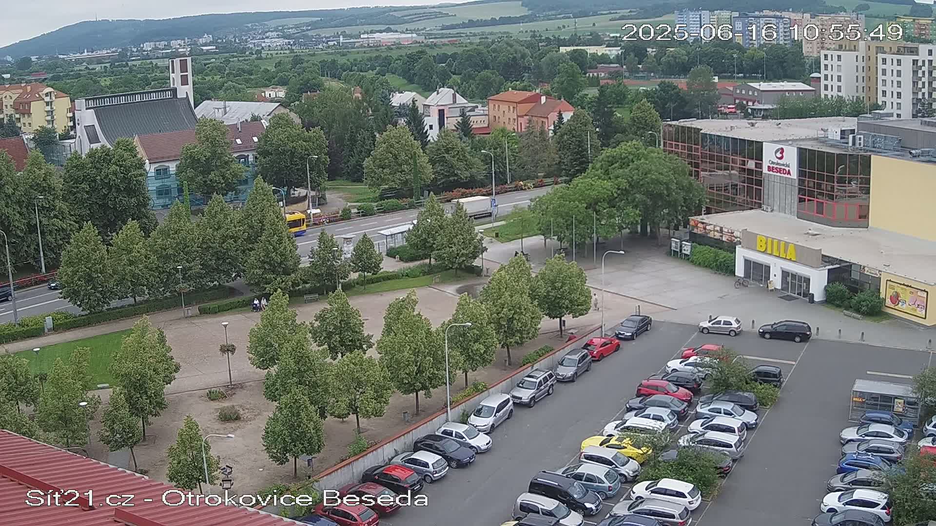 An overcast day reveals a town square with a church, a large parking lot filled with cars, and a Billa supermarket, all surrounded by lush green trees and buildings.