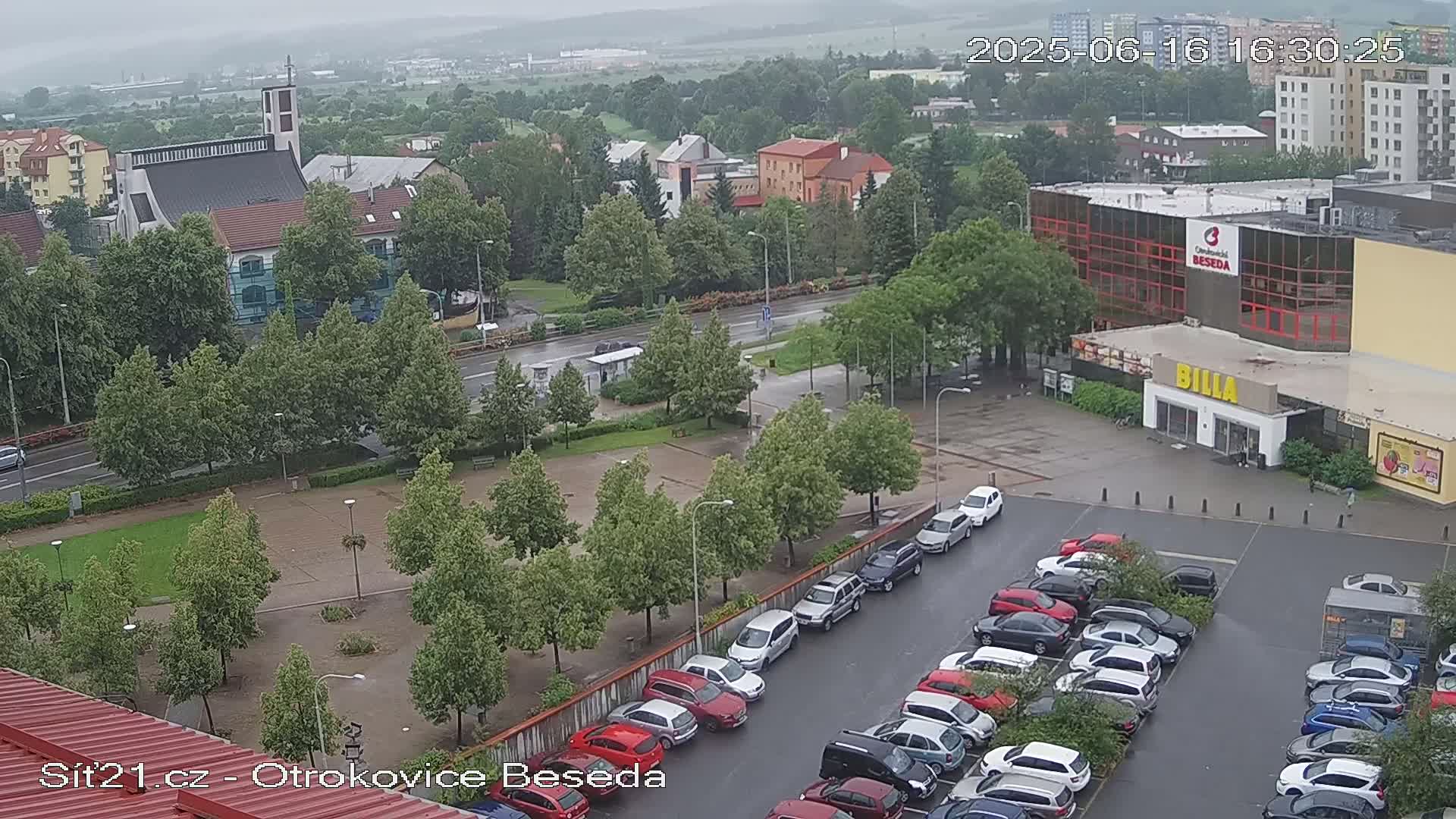 A high-angle, long shot of a town square on a rainy day shows a church, buildings, a parking lot full of cars, and many green trees.