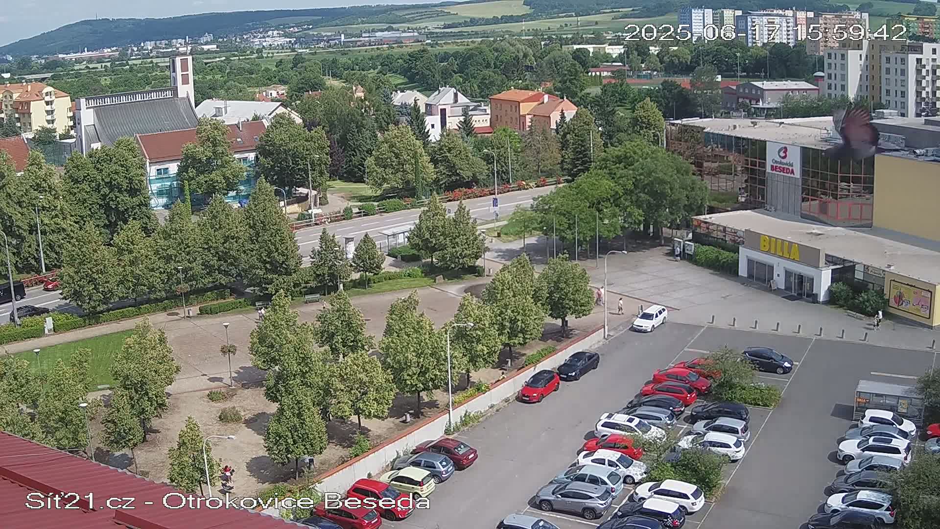 A high-angle view of a town square with a church, buildings, and a parking lot filled with cars on a sunny day.