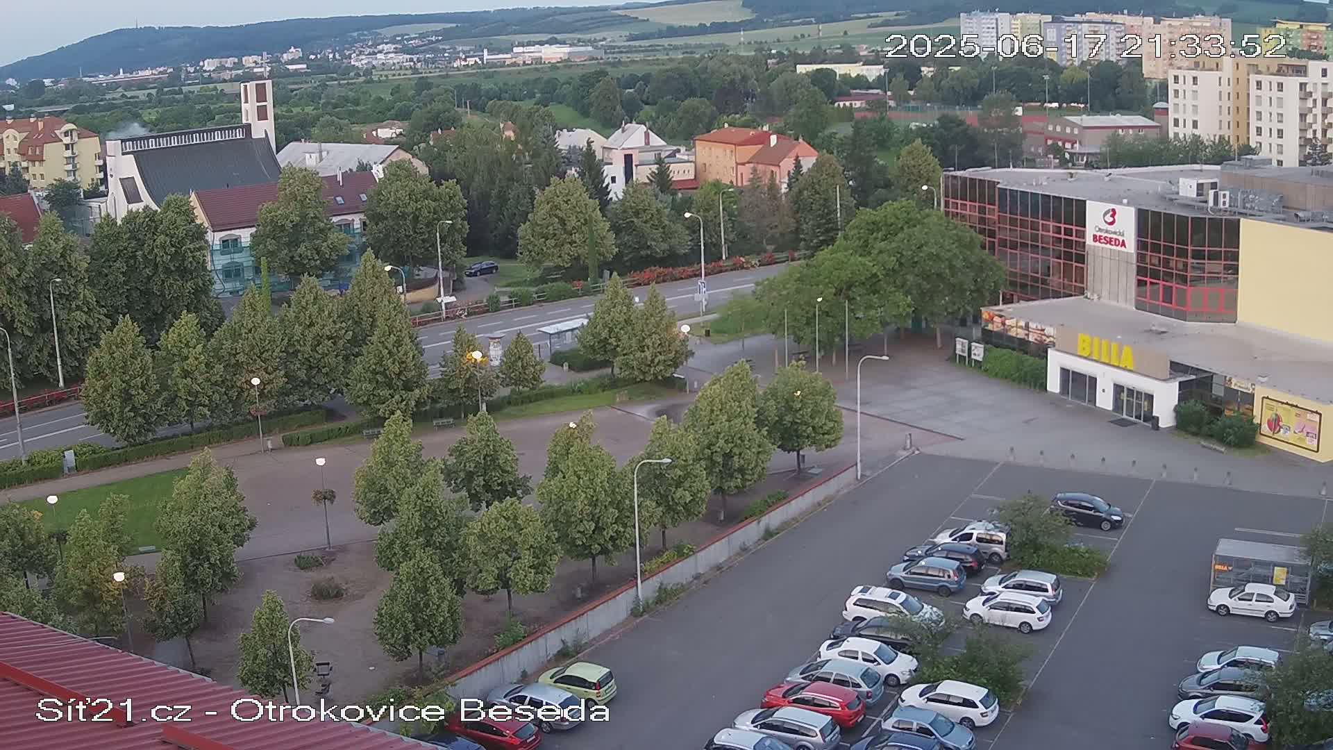 An aerial view of a town square with a church, a large building, and a parking lot filled with cars, under a partly cloudy sky.