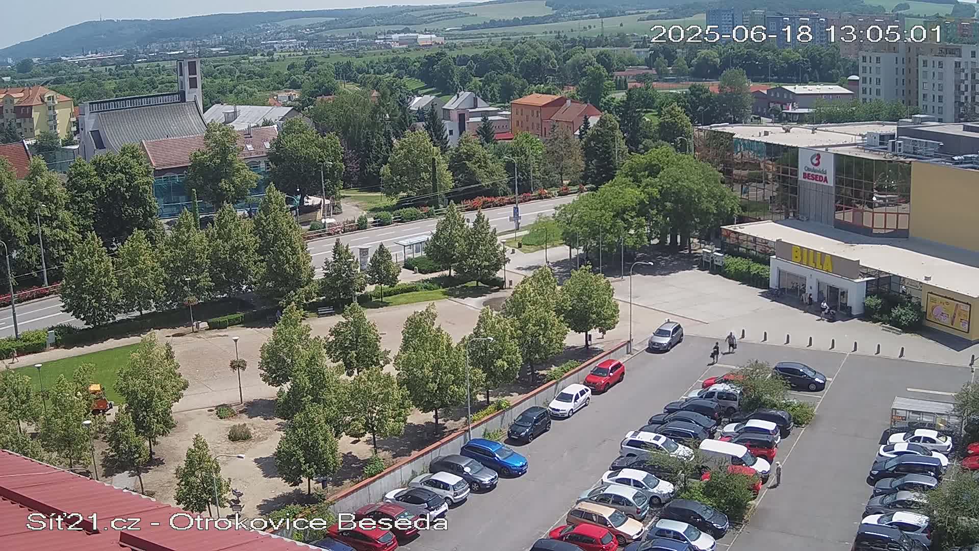 An aerial view of a town square with a church, shops, and a parking lot filled with cars on a sunny day.