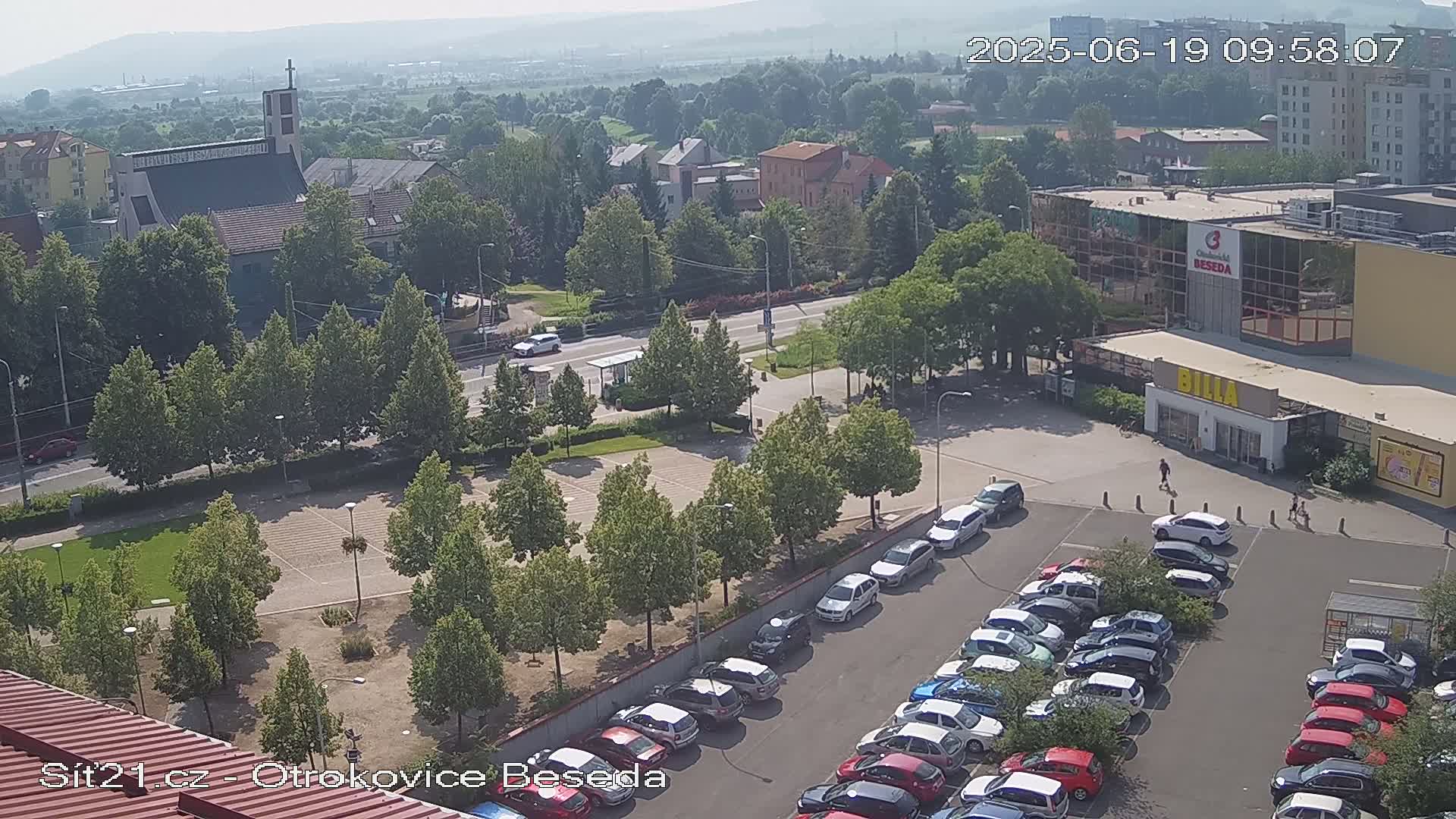 A high-angle view of a town square on a sunny day, showing a church, buildings, a parking lot filled with cars, and green spaces with trees.
